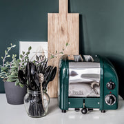 Dualit | 2 Slice Classic Toaster, Evergreen, displayed with a potted succulent and kitchen utensils, showcases its sleek design and versatile features for modern kitchens.