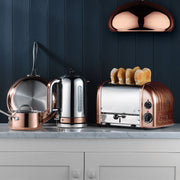 Dualit | 4 Slice Classic Toaster, Copper, toasting bread on a white marble countertop, surrounded by copper-accented kitchenware, under a copper pendant lamp.