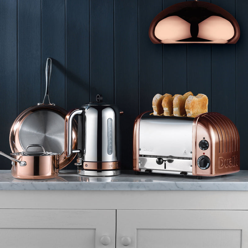 Dualit | 4 Slice Classic Toaster, Copper, toasting bread on a white marble countertop, surrounded by copper-accented kitchenware, under a copper pendant lamp.