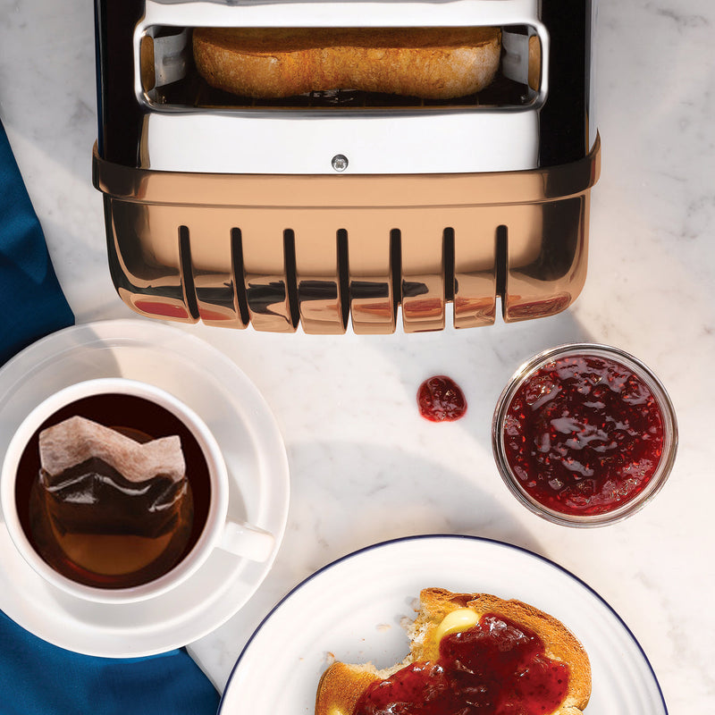 Dualit | 4 Slice Classic Toaster, Copper, with toast popping up, alongside tea, jam, and buttered toast on a marble surface, embodying culinary craftsmanship.