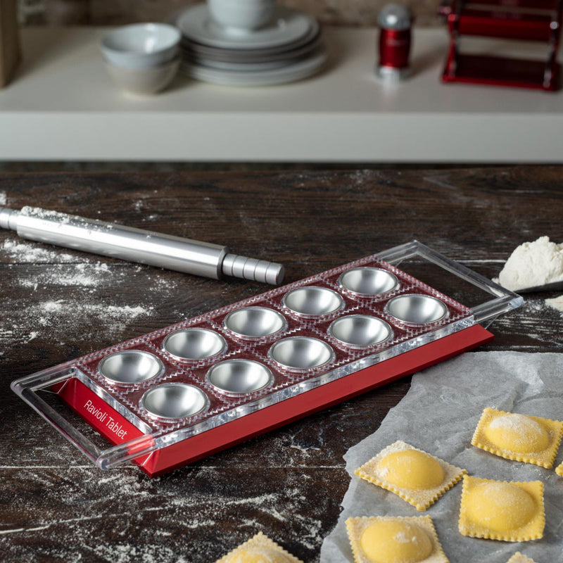 Marcato | Ravioli Tablet Pasta Makers, Powder Gray, with clear top and red base, forms fresh ravioli alongside scattered flour and a rolling pin on a wooden countertop.