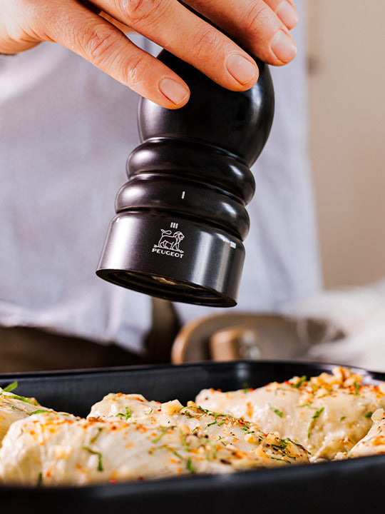Peugeot | Paris u'Select Pepper Mill, Satin Black in use, grinding fresh pepper over herb-seasoned chicken breasts, showcasing its adjustable grind feature.