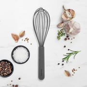 GreenPan | Premium Silicone Whisk, gray-handled, surrounded by cooking ingredients including garlic, salt, peppercorns, coriander, rosemary, and thyme on a white surface.