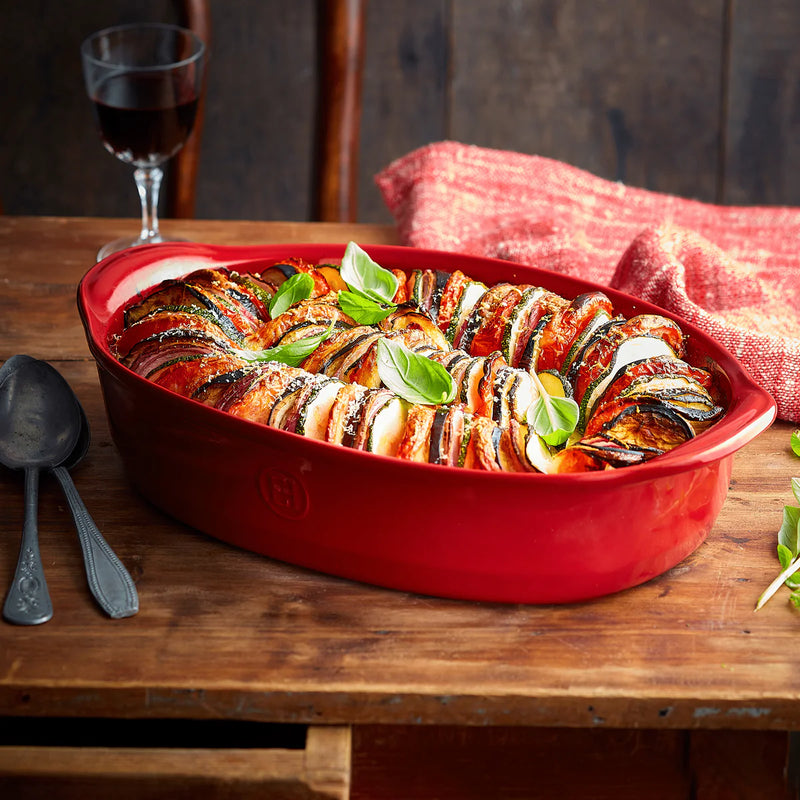 Emile Henry | The Right Baking Dish, Small Oval, Burgundy, filled with sliced zucchini, eggplant, and tomato, topped with cheese and basil, on a wooden table.