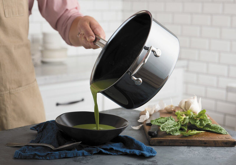 Hestan NanoBond 2qt Saucepan pours vibrant green soup into a black bowl, showcasing its precision pouring and ergonomic design amidst fresh kitchen ingredients.