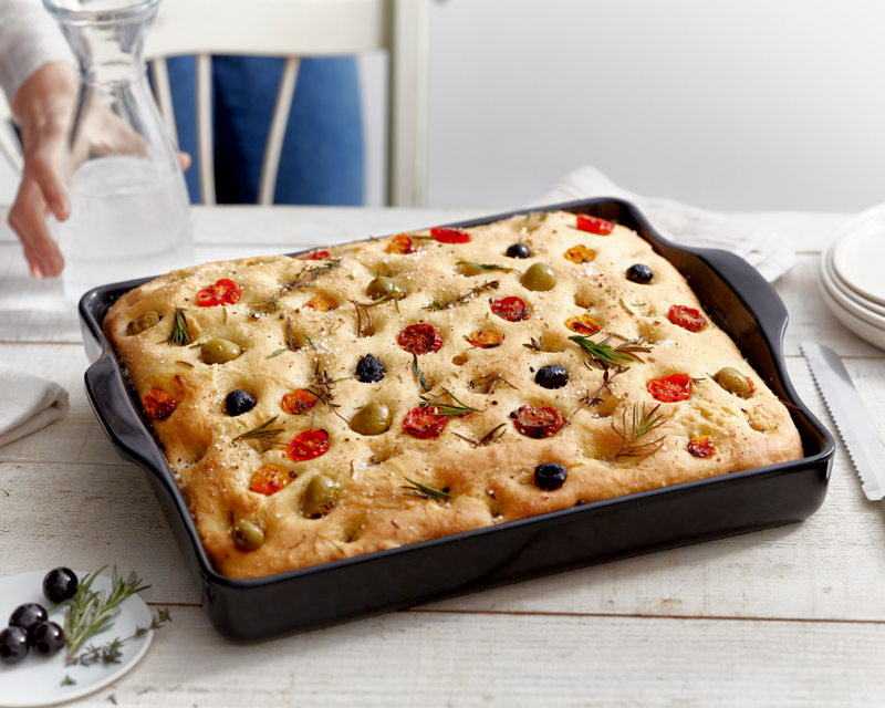 Emile Henry Bread Box, Truffle, with freshly baked focaccia in a black pan, featuring olives, tomatoes, and herbs. A glass carafe and plates are in the background.