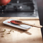 Emile Henry | Spoon Rest, Burgundy holds a wooden spoon with red sauce on a wooden countertop, ideal for tidy meal prep.