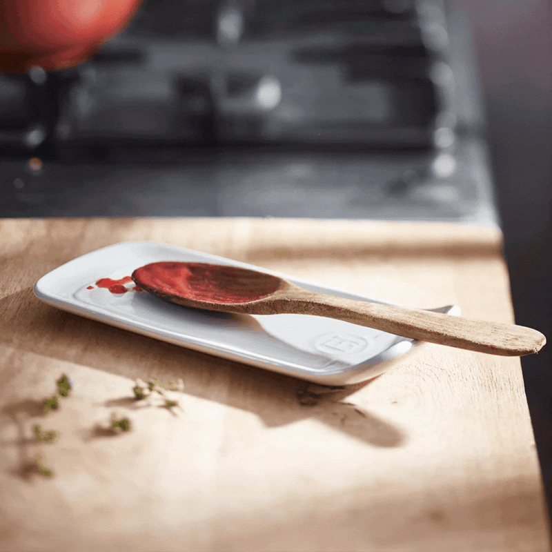Emile Henry | Spoon Rest, Burgundy holds a wooden spoon with red sauce on a wooden countertop, ideal for tidy meal prep.