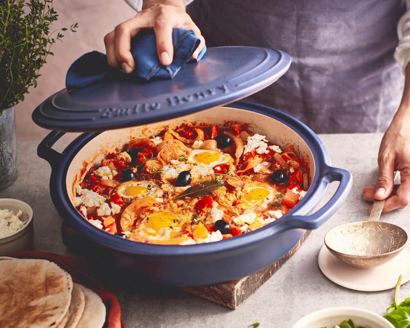 Emile Henry Garlic Pot, Burgundy, showcased with shakshuka in a blue Dutch oven, highlighting its ventilation design for fresh garlic storage.