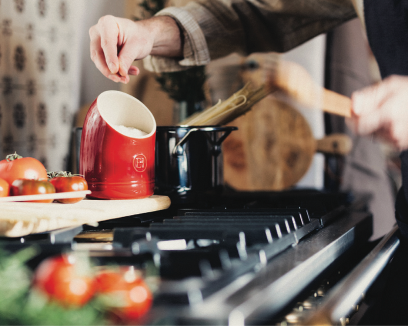 A hand sprinkles salt while cooking pasta beside fresh tomatoes, showcasing the Emile Henry | Focaccia + Detroit Pizza Pan, ideal for crispy, chewy Detroit-style pizza.