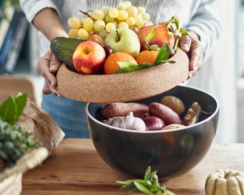 Emile Henry Deep Flan & Tart Dish on tabletop with two-tiered serving bowl of fruits and vegetables; hands lift cork-textured tray.