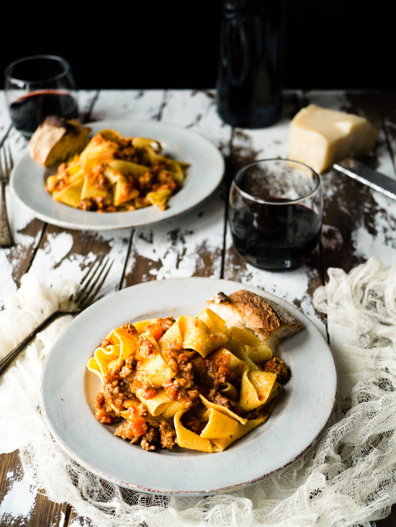 Nicola's Marketplace - Pappardelle served with meaty ragù, crusty bread, red wine, and cheese on a rustic table, showcasing premium, locally sourced ingredients.