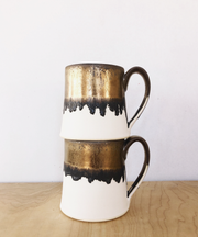 Jessica Wertz Ceramics - Gold + White Mug: Two handmade ceramic mugs, white with metallic bronze-gold glaze, stacked on a wooden surface, showcasing unique drip patterns and glossy interiors.