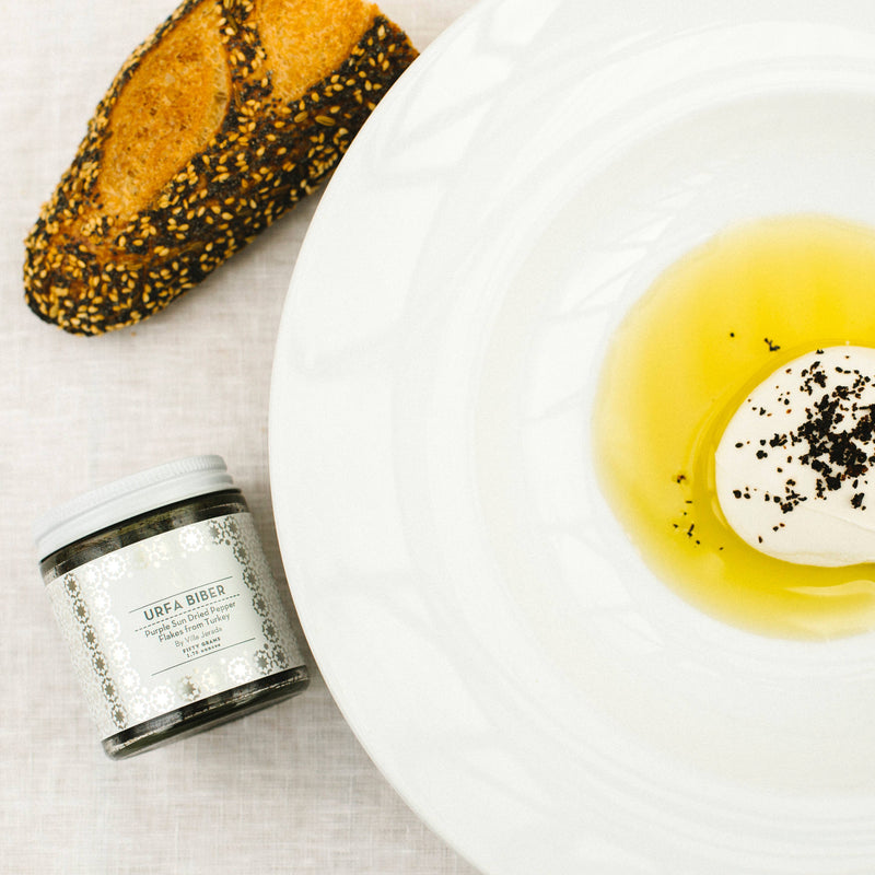 Villa Jerada - Urfa Biber - 50g jar beside seeded bread and cheese in olive oil, showcasing its rich, complex pepper flakes.