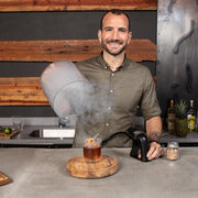 Man showcasing the Zwiesel Fortessa | Crafthouse Signature Smoke Cloche Set, featuring a smoked cocktail with citrus garnish, on a kitchen bar with tools and ingredients.