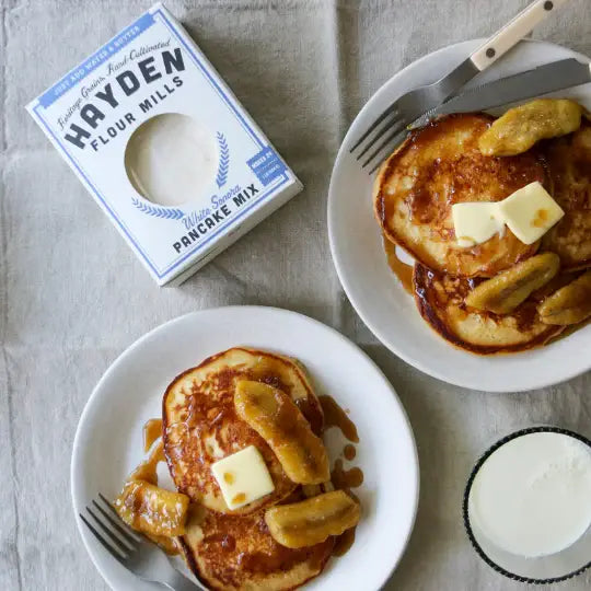 Hayden Flour Mills - White Sonora Pancake Mix with pancakes topped with butter, bananas, and syrup, accompanied by milk. Perfect for delicious, easy breakfast preparation.