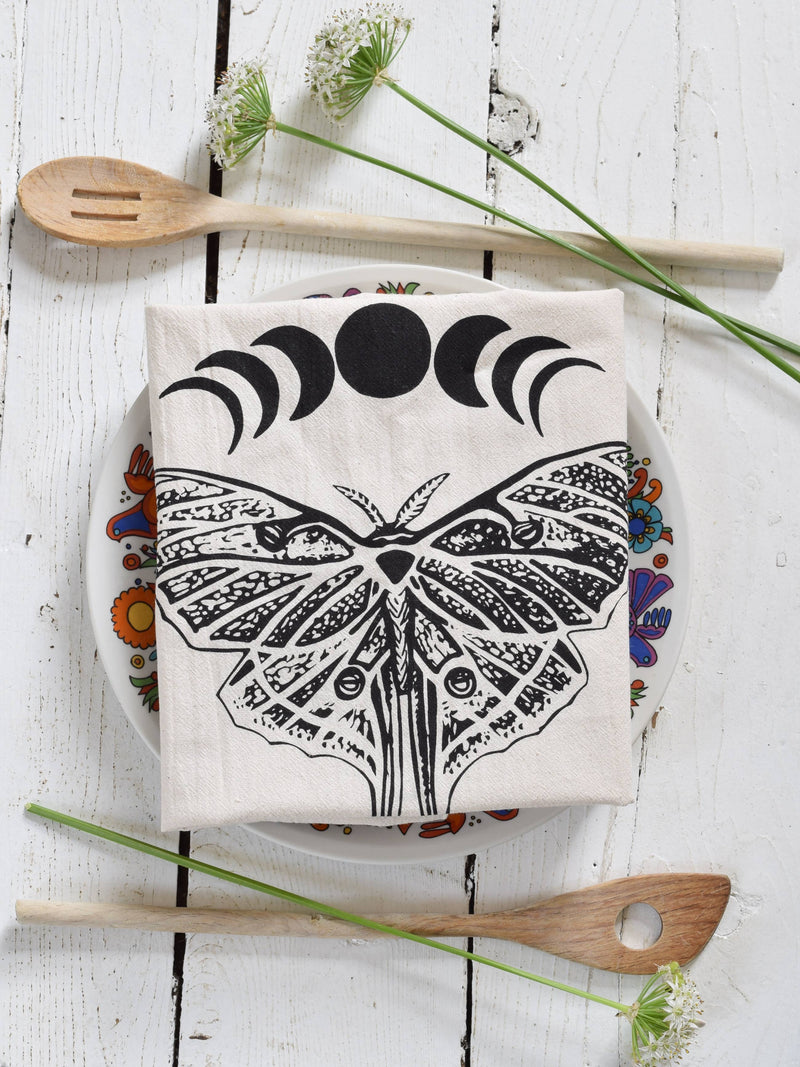 Hearth and Harrow - Organic Cotton Luna Moth Tea Towel with moon phases, displayed on a floral plate, accompanied by wooden spoons and delicate white flowers.