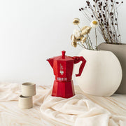 Bialetti | Red Moka Express Exclusive 3 Cup centered with vintage design, silicone gasket, and glossy finish, accompanied by ceramic cups and vases on a draped surface.