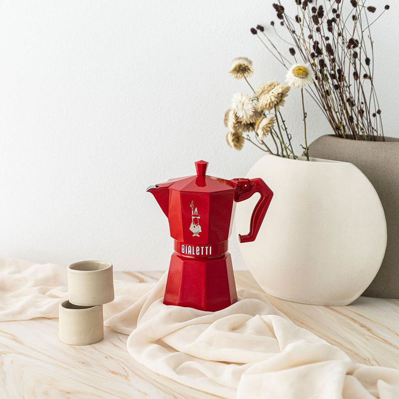 Bialetti | Red Moka Express Exclusive 3 Cup centered with vintage design, silicone gasket, and glossy finish, accompanied by ceramic cups and vases on a draped surface.