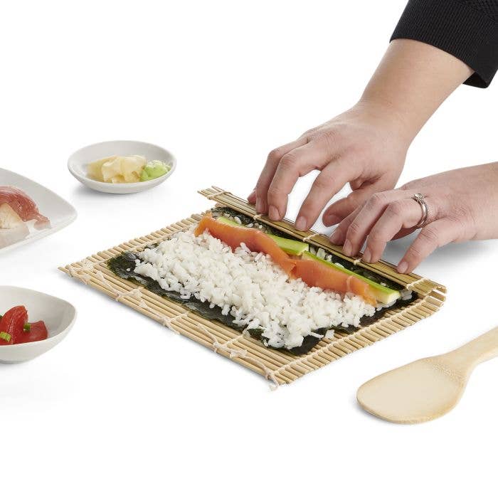 Hands roll sushi using HIC - Harold Import Co. - Helen's Asian Kitchen Sushi Mat With Paddle, shaping nori, rice, salmon, cucumber, and avocado into a roll.
