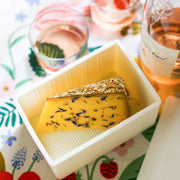 CapaBunga® Cheese Vault® in use, featuring a cheese wedge with blue petals and herbs, alongside rosé wine on a patterned tablecloth.