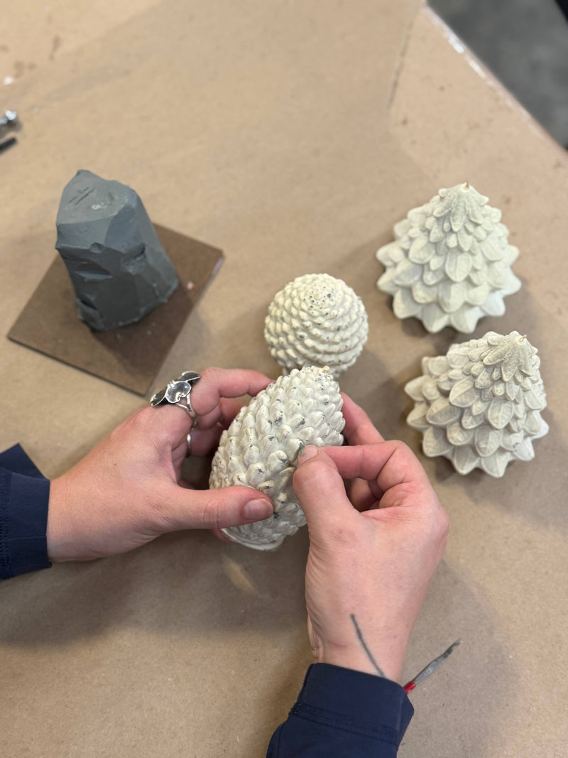 Hands refine a sculpted beeswax pine cone candle on a worktable, showcasing Mo&Co Home's exclusive design in two sizes: large (H: 4, D: 2 3/4).