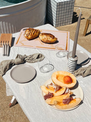 Outdoor table with The MK Free Board: Ocean holding grilled chicken, alongside cantaloupe with prosciutto, stoneware plates, and a candle, showcasing Athens Cooks' curated kitchen essentials.