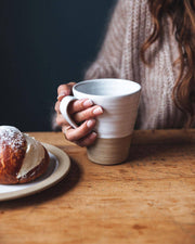 Hand holding Farmhouse Pottery - Tall Silo Mug beside a cream-filled brioche on a wooden tabletop, showcasing the mug's handcrafted, two-tone design and generous size.