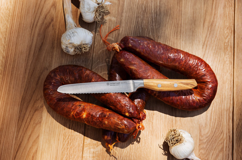 Wüsthof Amici 5 Serrated Utility Knife placed beside assorted sausages, showcasing its precision and versatility for culinary tasks.