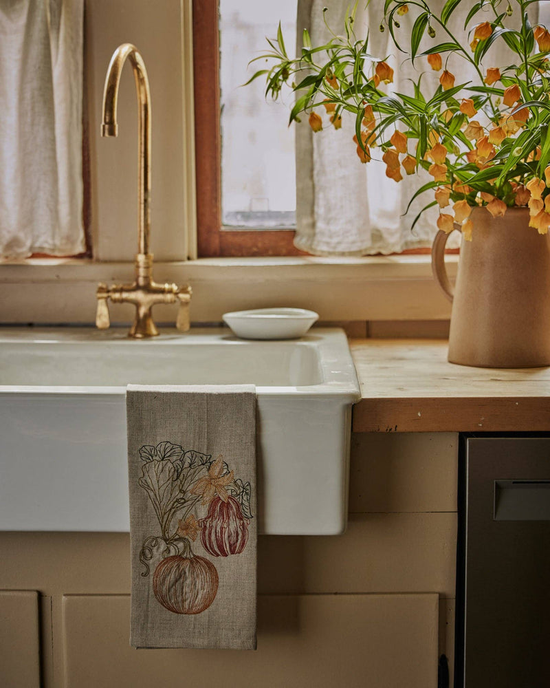 Cozy kitchen with Coral & Tusk - Pumpkin Vine Tea Towel on a farmhouse sink, featuring embroidered pumpkins and foliage, alongside a vintage brass faucet and ceramic pitcher.