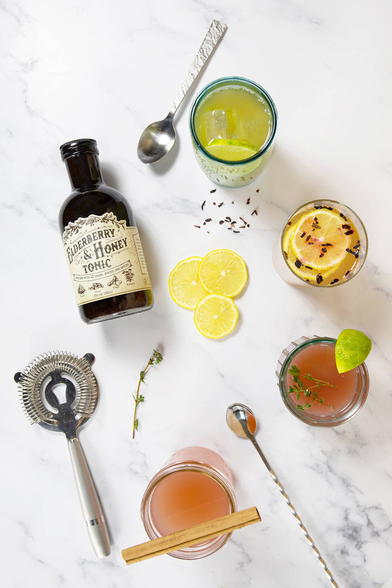 Roots And Leaves - Elderberry & Honey Tonic, 16 oz, in a glass bottle next to a glass of pink liquid with a lime wedge.