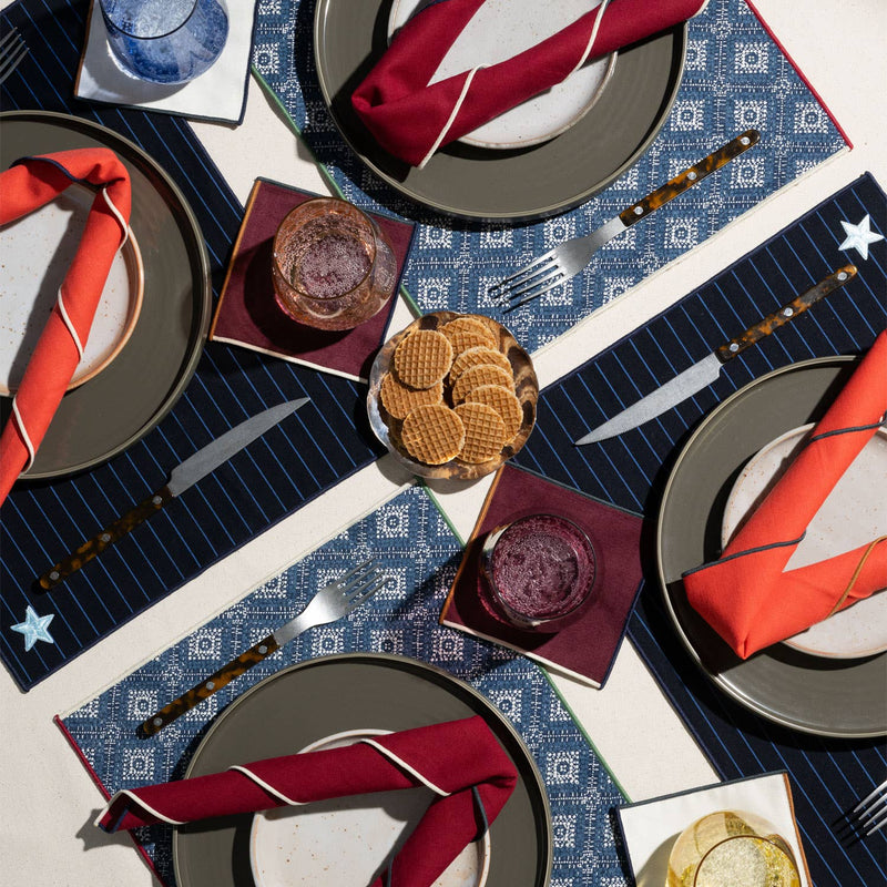 ATELIER SAUCIER - Clubhouse Cocktail Napkins: Set of 4, layered on a stylish table setting with patterned placemats, showcasing elegant tortoiseshell cutlery and vibrant coasters.