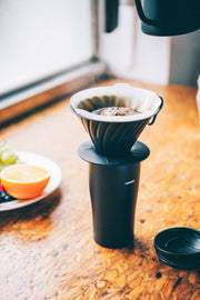 HARIO USA - V60 Metal Dripper: Silver on travel mug with coffee brewing; alongside fruit plate and pouring kettle.