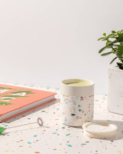 W&P - Reusable Ceramic Mug: Terrazzo Charcoal, 16 oz, with a green drink, beside a plant in a pot and a book.