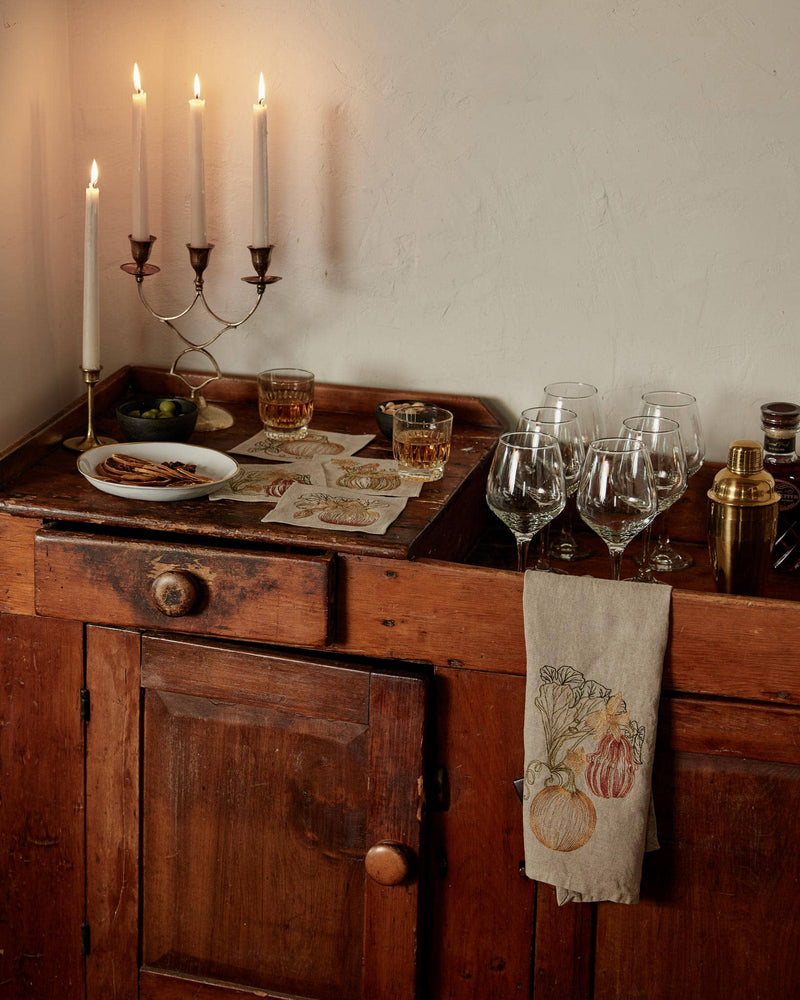 Coral & Tusk - Pumpkin Vine Tea Towel draped on a rustic bar cart with candelabra, glasses, snacks, creating a cozy autumnal entertaining setup.