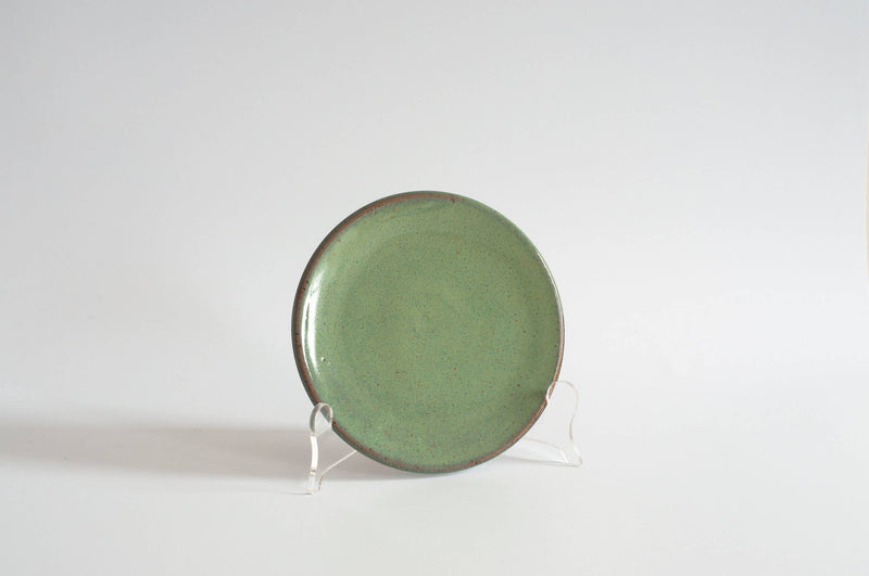 RachaelPots - Dessert Plate: Sage, a green speckled ceramic plate with a slightly upturned lip, shown upright on a clear stand.