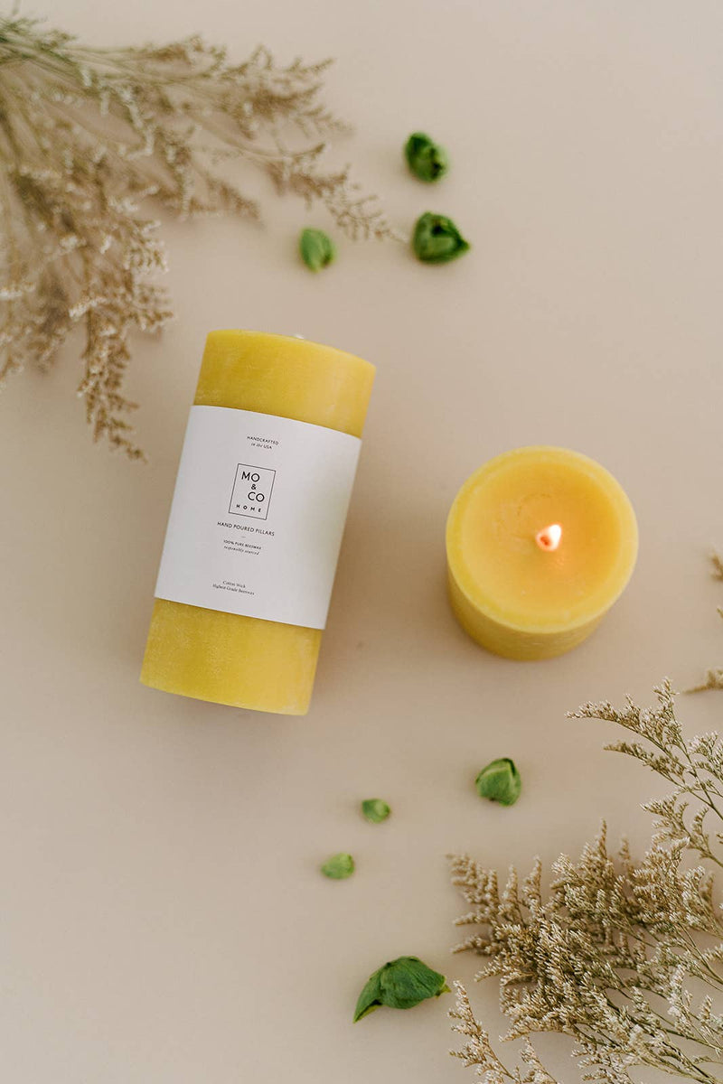 Mo&Co Home - 100% Beeswax Pillar Candles | Natural Gold: 3x4 Inch, hand-poured, eco-conscious, displayed on a white surface with green foliage.