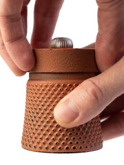 Hands adjusting the Peugeot | Bali Cast Iron Pepper Mill, Orange, showcasing its textured cylindrical body and adjustable top knob for setting grind coarseness.