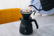 HARIO USA - V60 Metal Dripper: Silver, elegantly pouring over coffee, showcasing precision brewing with a black gooseneck kettle into a matching carafe.