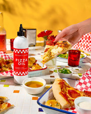 Brightland Pizza Oil bottle surrounded by a vibrant pizza party setup with cheese pizza slice, pepperoni slices, dips, garlic knots, and red tumblers on a colorful table.