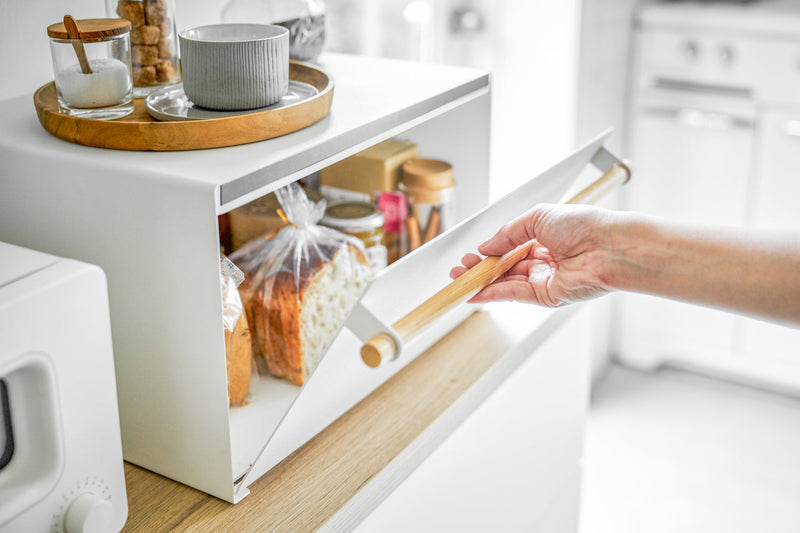 Yamazaki Home | Simple Steel Bread Box, White + Wood, on a countertop with bread inside, jars atop, and a hand opening its front flap.
