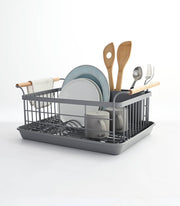 Yamazaki Home | Steel + Wood Dish Rack with Drainer Spout, featuring upright plate slots, utensil holder, and ash wood handles, showcased with kitchenware and a towel on the handle.