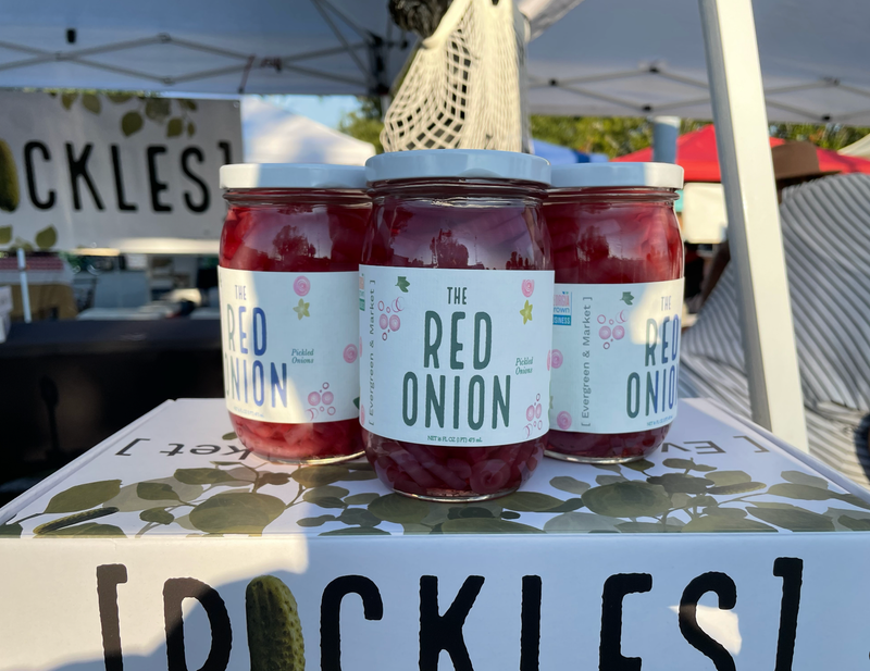 Three jars of PICKLES by Evergreen and Market - The Red Onion, showcased on a market stall, highlighting their unique pickled red onion flavor.
