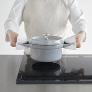 A person uses Yamazaki Home | Magnetic Pot Holders, Silicone, Black to lift a grey enameled pot from a black induction stove, showcasing their heat-resistant grip.