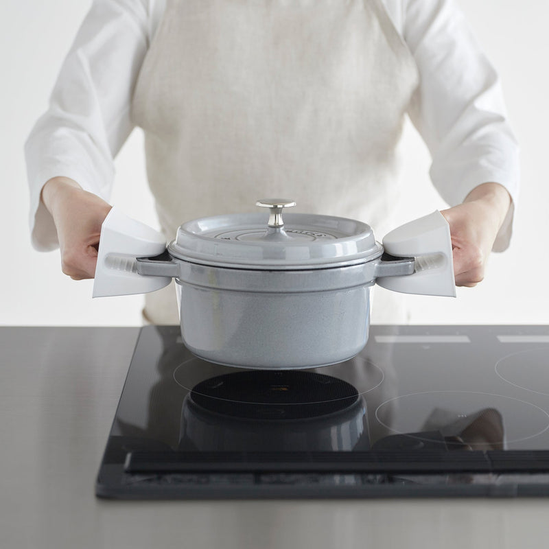 A person uses Yamazaki Home | Magnetic Pot Holders, Silicone, Black to lift a grey enameled pot from a black induction stove, showcasing their heat-resistant grip.
