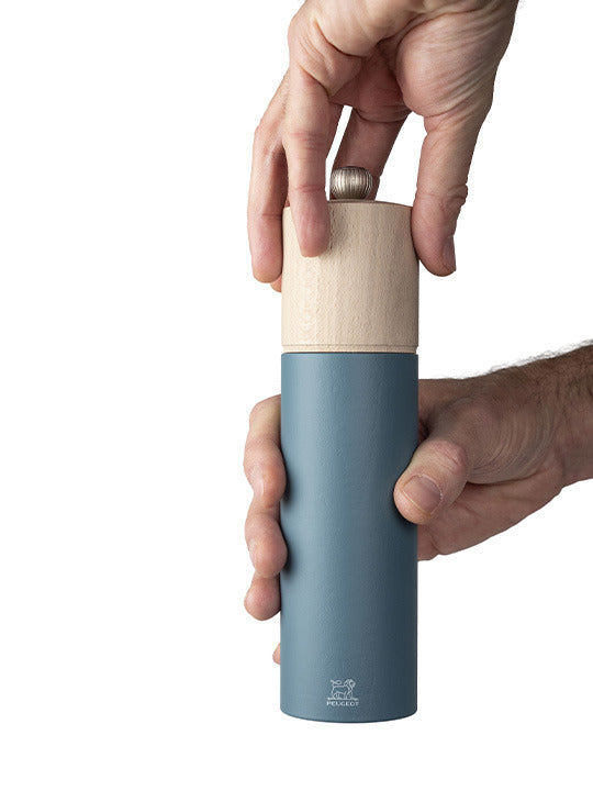 Peugeot | Boreal Pepper Mill, Celestial Blue, held by two hands, showcasing its matte teal body and light wood top with a small metal knob.
