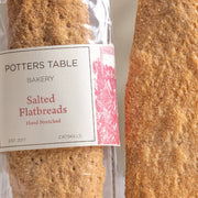 Potters Table - Salted Olive Oil Flatbreads, a packaged bakery item showing hand-stretched flatbreads; one in clear wrapper, one lying beside it, ideal for snacking.