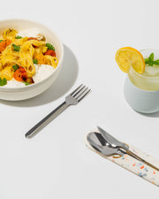 Stainless steel utensils set with a fork, spoon, and silicone carrying case, displayed next to a bowl of pasta and a drink.