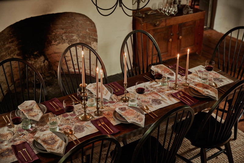 Rustic dining table with Coral & Tusk - Sunflower and Wheat Dinner Napkins, floral runner, brass candlesticks, gold-tone cutlery, and wine glasses, set in a cozy, old-world style room.