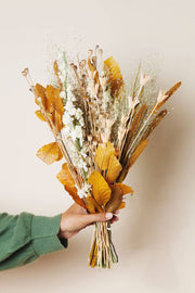Hand in a green sleeve holds the Idlewild Floral Co. - Golden Fall Market Bouquet, featuring dried botanicals with golden leaves and seed pods against a light background.